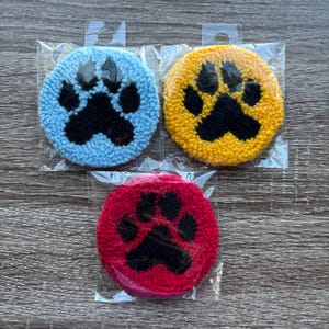 May include: Three colorful, round paw print coasters. One is light blue, one is yellow, and one is red. Each coaster has a black paw print design and is packaged in a clear plastic sleeve.