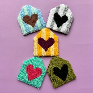 May include: Five colorful, hand-stitched felt house-shaped coasters with different colored hearts in the center. The coasters are in shades of blue, yellow, green, gray, and brown.