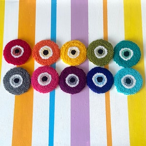May include: Ten round, colorful, crocheted coasters with a white evil eye design. The coasters are arranged in a row on a striped background.