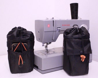 May include: Two black drawstring bags with orange drawstrings, one with a cell phone inside, are placed on a white surface in front of a gray Singer sewing machine.