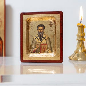 Op de afbeelding: Een religieus icoon met een bebaarde man in een gewaad, die een boek vasthoudt. Het icoon is gevat in een bladgouden lijst met een rode rand. Een brandende kaars in een gouden houder en een boeket bloemen staan op de achtergrond.