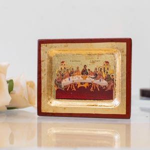 May include: A small, framed icon depicting the Last Supper. The icon is painted in a traditional style with gold accents and a red background. The frame is made of wood and has a dark red finish.