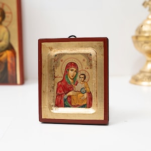 Pode incluir: Um ícone religioso retratando a Virgem Maria e o menino, emoldurado em uma moldura de madeira vermelha escura com detalhes dourados. O ícone apresenta obras de arte detalhadas com tons de vermelho, dourado e azul. Um segundo ícone é visível ao fundo.