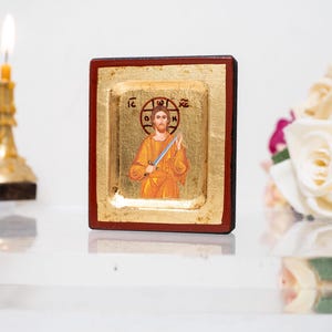 Könnte beinhalten: Religiöse Ikone mit einer Figur in einem goldenen Gewand, die ein Schwert hält, vor einem goldenen Hintergrund mit einem dunklen Holzrahmen. Eine brennende Kerze und weiße Rosen befinden sich im Hintergrund.