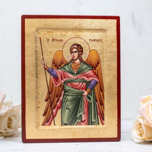 May include: A religious icon depicting a winged figure with a halo and staff. The figure wears green, red, and purple robes, with gold detailing. The icon is set in a wooden frame with a gold leaf background. The text "O APXON" and "RAPAHA" is visible.
