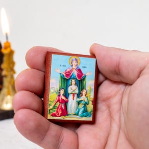May include: A small wooden icon with a colorful image of the Virgin Mary and three children. The icon is held in a hand against a white background.