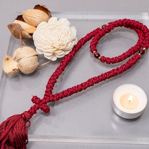 May include: A red prayer rope with a tassel and a cross charm, a small white candle, and dried flowers on a clear glass surface.
