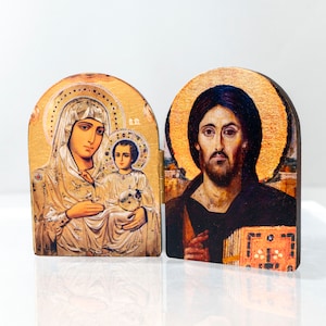 Puede incluir: Un conjunto de iconos plegables de madera con dos paneles. El panel izquierdo representa a la Virgen María sosteniendo al niño Jesús. El panel derecho representa a Jesucristo con un libro en la mano. Ambos paneles tienen un fondo dorado.