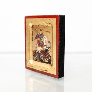 May include: A small, framed icon depicting Saint George slaying a dragon. The icon is painted in vibrant colors and features gold accents. The frame is made of wood and has a red and black finish.