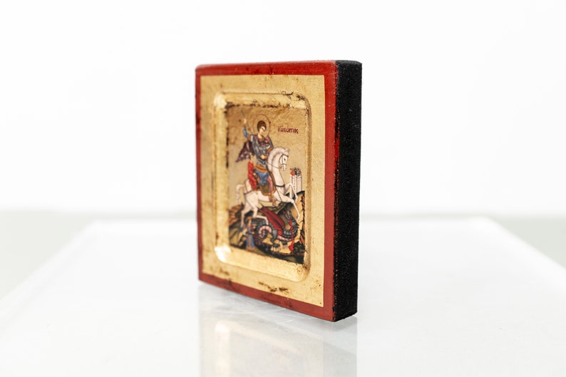 May include: A small, framed icon depicting Saint George slaying a dragon. The icon is painted in vibrant colors and features gold accents. The frame is made of wood and has a red and black finish.