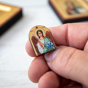 May include: A small, arched wooden icon depicting a religious figure with a halo, wings, and a shield. The icon is held between fingers, with two other icons in the background. The artwork features a detailed illustration with vibrant colors and gold accents.