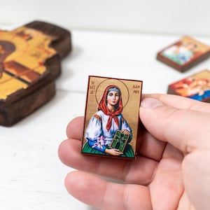 May include: A small, rectangular religious icon featuring a woman with a halo, wearing a red scarf and holding a book. The icon is held in a hand, with other similar icons and a wooden cross in the background.