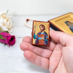 Op de afbeelding: Een klein houten icoon met een geschilderd beeld van een man die een kruis vasthoudt. Het icoon bevindt zich in een hand, tegen een witte achtergrond.