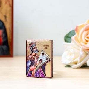 Può includere: Una piccola icona rettangolare in legno raffigurante un uomo barbuto con corona e vesti viola decorate, che tiene in mano uno strumento a corda triangolare. Lo sfondo è dorato e vicino alla testa della figura è visibile del testo in greco.