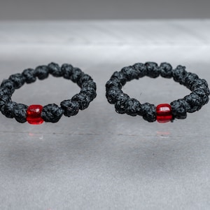 Puede incluir: Dos pulseras de oración de nudo negro con una sola cuenta roja en cada una.