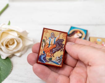 May include: A small, rectangular religious icon featuring a saint on horseback, with gold and red accents. The icon is held in a hand, with other similar icons visible in the background. A white rose is in the background.