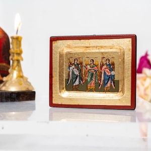 May include: A small, framed icon with a gold background and red border. The icon depicts three angels with wings, each holding a staff and wearing a robe. The angels are standing in a row, facing forward.
