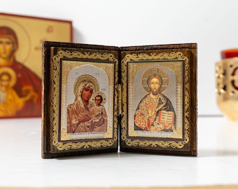 Könnte beinhalten: Ein hölzernes, aufklappbares Ikon mit zwei religiösen Bildern. Die linke Tafel zeigt Maria und Jesus, während die rechte Tafel Jesus mit einem Buch zeigt. Beide Tafeln haben goldene Akzente und sind in einem braunen Holzrahmen gefasst.