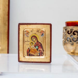 May include: A small, wooden framed icon depicting the Virgin Mary holding a baby Jesus. The icon has a gold background and is surrounded by a red frame.