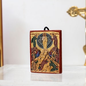 Pode incluir: Um pequeno &iacute;cone de madeira que representa a Ascens&atilde;o de Jesus Cristo. O &iacute;cone &eacute; pintado em cores vibrantes e apresenta Jesus ascendendo ao c&eacute;u com anjos e disc&iacute;pulos abaixo. O &iacute;cone &eacute; emoldurado em uma moldura de madeira vermelha.