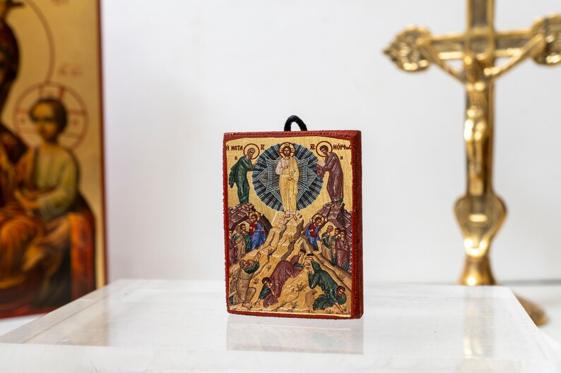 Pode incluir: Um pequeno &iacute;cone de madeira que representa a Ascens&atilde;o de Jesus Cristo. O &iacute;cone &eacute; pintado em cores vibrantes e apresenta Jesus ascendendo ao c&eacute;u com anjos e disc&iacute;pulos abaixo. O &iacute;cone &eacute; emoldurado em uma moldura de madeira vermelha.