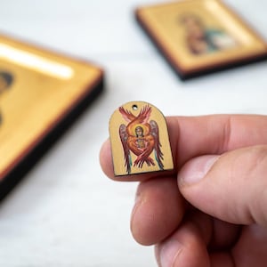 Peut inclure: Petite icône religieuse en bois représentant un ange aux ailes rouges, oranges et jaunes. L'icône est tenue dans une main, avec deux autres icônes en arrière-plan. L'icône a un trou en haut.