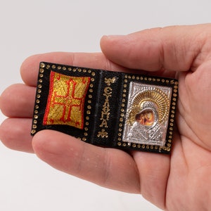 May include: A miniature religious icon in the shape of an open book. The black cover has gold detailing and a gold and red cross. The right side displays a silver icon with a gold halo. The item is held in a hand.