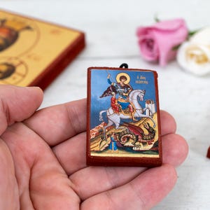 May include: A small, rectangular icon featuring a depiction of Saint George on horseback slaying a dragon. The icon has a blue background and is framed in red. Other icons and flowers are in the background.
