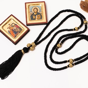 Könnte beinhalten: Schwarzes Gebetsseil mit goldenen Perlen und einem Quasten. Das Seil ist aufgerollt und auf einer weißen Oberfläche ausgelegt. Zwei kleine gerahmte religiöse Ikonen sind ebenfalls auf dem Bild zu sehen.