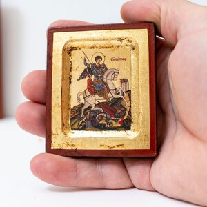 May include: A small wooden framed icon depicting Saint George slaying a dragon. The icon is painted in vibrant colors and features gold accents. The text "ά&gamma;&iota;&omicron;&sigmaf; &Gamma;&epsilon;ώ&rho;&gamma;&iota;&omicron;&sigmaf;" is written in Greek at the top of the icon.
