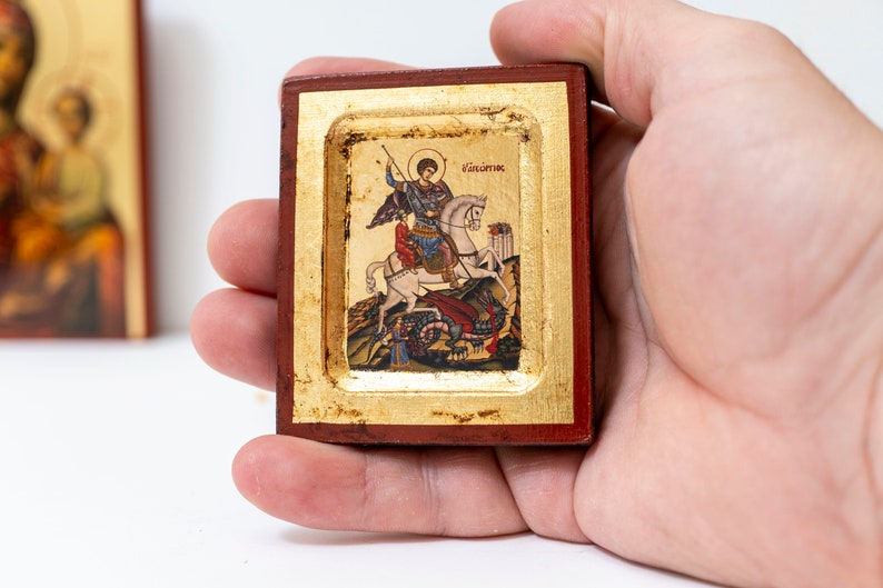 May include: A small wooden framed icon depicting Saint George slaying a dragon. The icon is painted in vibrant colors and features gold accents. The text "ά&gamma;&iota;&omicron;&sigmaf; &Gamma;&epsilon;ώ&rho;&gamma;&iota;&omicron;&sigmaf;" is written in Greek at the top of the icon.