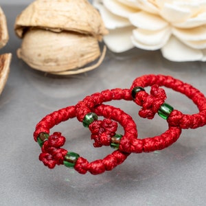 May include: Three red string bracelets with green beads. The bracelets are knotted together and have a cross design.