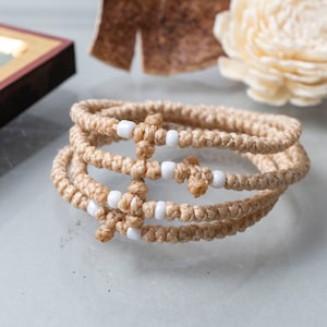May include: A set of three beige prayer ropes with white beads and a cross charm. The ropes are knotted and braided together.