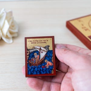 May include: A small, rectangular religious icon with a red border. The icon shows a boat with figures on blue water, with text at the top. A larger icon with a depiction of Jesus is in the background.
