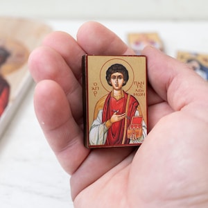 Può includere: Una piccola icona rettangolare raffigurante una figura religiosa con un'aureola, vestita con abiti rossi e bianchi, che tiene una piccola scatola. L'icona ha dettagli dorati e testo greco. Altre immagini religiose sono visibili sullo sfondo.
