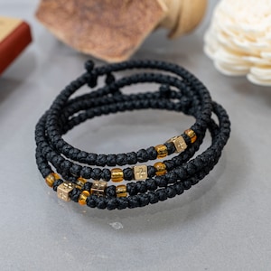 Könnte beinhalten: Ein schwarzes Gebetsrosenarmband mit goldenen Perlen und Kreuzen. Das Armband ist aus geflochtener Schnur gefertigt und hat am Ende einen Knoten.
