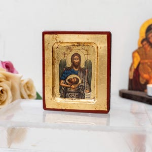 Op de afbeelding: Een klein, ingelijst icoon dat een religieus figuur voorstelt dat een hoofd in zijn armen houdt. Het icoon staat op een wit oppervlak met een kaars en een groter icoon op de achtergrond.