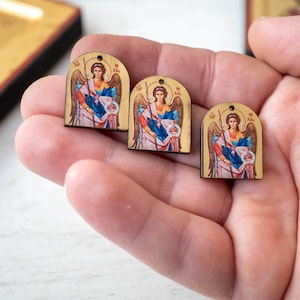 Op de afbeelding: Drie houten hangers met een religieus icoon van een engel met vleugels, die een staf vasthoudt. De engel is afgebeeld in kleurrijke gewaden met een halo. De hangers worden in een hand gehouden, met een groter icoon op de achtergrond.