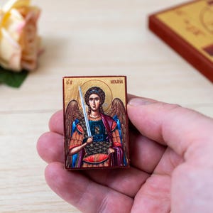 May include: A small rectangular wooden icon featuring a painted depiction of an angel with wings, holding a sword. The figure has a golden halo and wears armour with red and blue garments. The text "MIXAIL" is printed in the upper right corner.