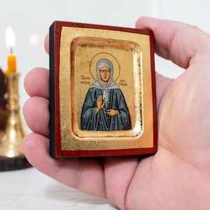 May include: A small wooden icon with a gold frame depicting a woman in a blue robe with a gold halo. The icon has Greek text on it that reads "Άγιος Νικόλαος" and "Παναγία".