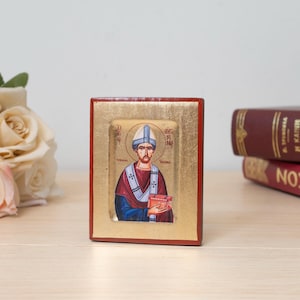 Könnte beinhalten: Eine rechteckige religiöse Holzikone mit einem zentralen Gemälde einer bärtigen Figur in weißer Mitra und rot-blauem Gewand, die eine kleine rote Schachtel hält. Die Ikone hat einen Blattgoldrand und einen dunkelroten Rahmen mit kyrillischem Text.