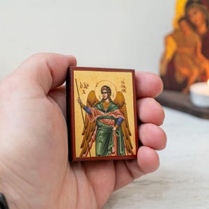 Può includere: Una piccola icona in legno con un'immagine dipinta di un angelo che tiene una lancia. L'icona ha uno sfondo dorato e un bordo rosso. L'angelo indossa una veste verde e ha le ali. L'icona è tenuta nella mano di una persona.