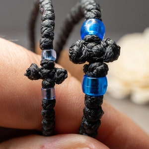 Op de afbeelding: Twee zwarte gevlochten armbanden met blauwe en heldere kralen en een kruisbedel.