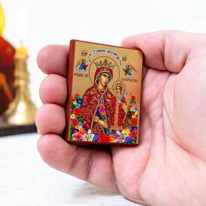 Può includere: Una piccola icona in legno che raffigura la Vergine Maria e il bambino Gesù. L'icona è dipinta in oro, rosso e blu con accenti floreali. L'icona è tenuta in una mano.