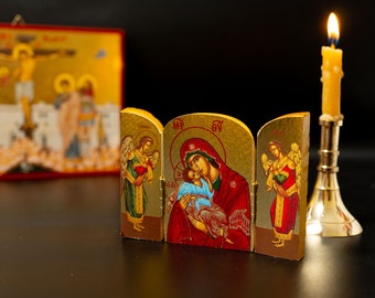 Könnte beinhalten: Ein goldfarbenes, dreiteiliges religiöses Icon, das die Jungfrau Maria mit dem Jesuskind zeigt, flankiert von Engeln. Eine brennende Kerze in einem goldenen Halter und ein gerahmtes religiöses Bild befinden sich im Hintergrund.