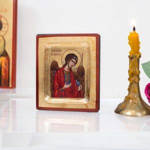 以下が含まれることがあります： 金色の背景に翼と青いローブを着た天使を描いた、小さな木製の額縁のアイコン。アイコンは、点灯したろうそくと花のある白い表面の上に置かれています。