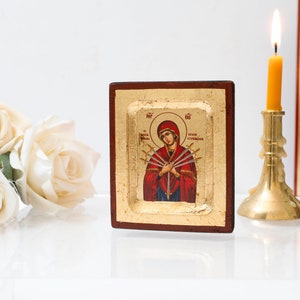 May include: A small wooden icon with a gold background depicting a woman in red and blue robes with seven swords piercing her heart. The icon is framed in a dark wood frame and is sitting on a white surface with white roses in the foreground. A lit candle is in a brass candlestick to the right of the icon.