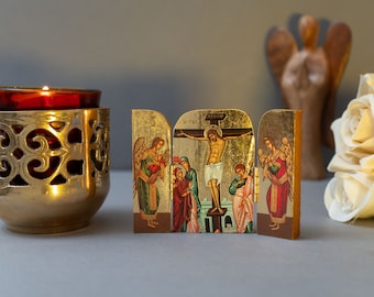 May include: A gold-colored candle holder with an intricate cut-out design holds a lit red candle. A three-panel icon depicting a religious scene with angels and figures is in the center. A wooden angel statue and a white rose are in the background.