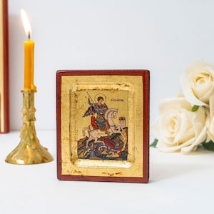 May include: A small, framed icon depicting Saint George slaying a dragon. The icon is painted in a traditional style with bright colors and gold accents. The frame is made of wood and has a red and gold finish.