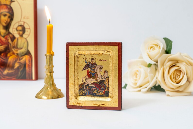 May include: A small, framed icon depicting Saint George slaying a dragon. The icon is painted in a traditional style with bright colors and gold accents. The frame is made of wood and has a red and gold finish.